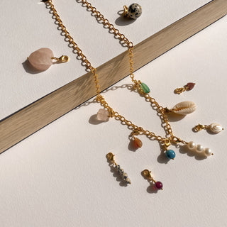 Collier charms personnalisable Youm posé à plat, lumière douce, mettant en valeur la chaîne dorée et le choix de charms en pierre et perle.
