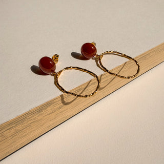 Boucles d’oreilles Luke ovale pendante dorée avec cornaline Youm, finition or fin 24 carats, pose pour montrer le détail de la dorure et de la pierre.